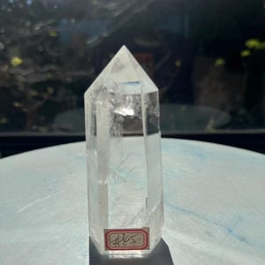 May include: A clear, hexagonal quartz crystal point with a pointed top and a flat base. The crystal is transparent with visible internal inclusions and fractures. A small rectangular white sticker with a red border and the handwritten text #65 is attached to the lower front section of the crystal.