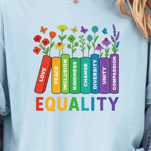 May include: Pale blue t-shirt with a vibrant graphic. The design shows a row of books with words such as "Love" and "Equality" and flowers. The word "Equality" is written in rainbow colours.