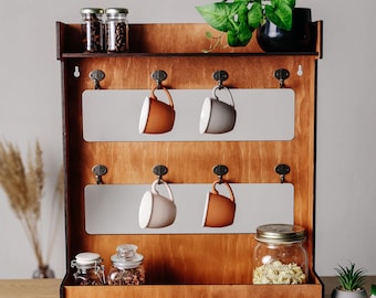 Può includere: Un ripiano a muro in legno con ganci per appendere tazze. Il ripiano ha due livelli e uno scaffale inferiore per riporre oggetti. Ci sono quattro tazze appese ai ganci, due marroni e due bianche. Ci sono anche tre barattoli di vetro sullo scaffale inferiore.
