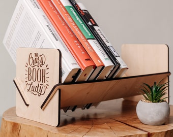 May include: A wooden book stand with the text "The Crazy Book Lady" engraved on the front. The stand is holding a stack of books with Russian titles. The stand is sitting on a wooden surface.