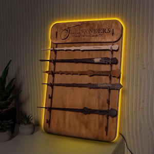 May include: A wooden wall-mounted display with the text "Ollivanders Makers of Fine Wands since 382 BC" and five wooden wands with different designs and names. The display is lit with a warm yellow light.