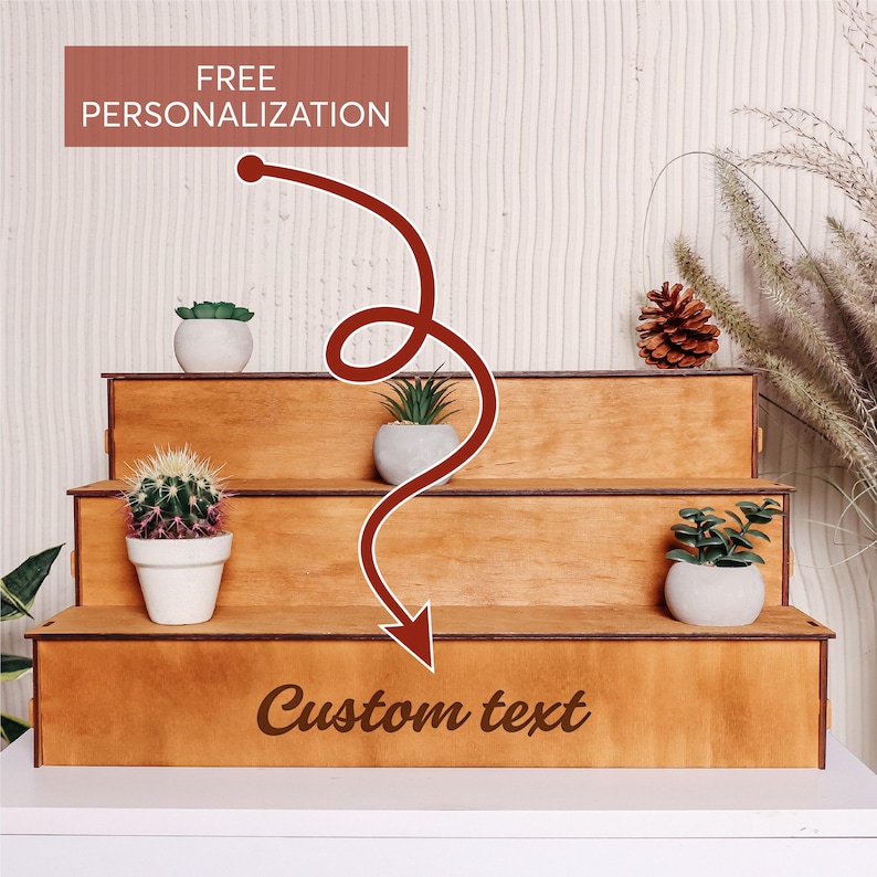 Pode incluir: Unidade de prateleira de madeira de tr&ecirc;s n&iacute;veis com acabamento em madeira natural. A unidade de prateleira &eacute; decorada com plantas em vasos e uma pinha. A prateleira inferior tem o texto "Custom text" gravado.