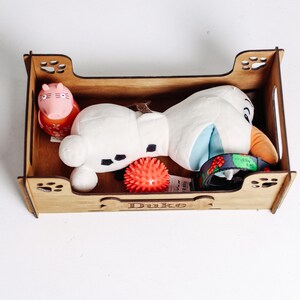 May include: A wooden dog bed with the name "Duke" engraved on the front. The bed contains a plush Olaf toy, a pink pig toy, and a red spiky ball.
