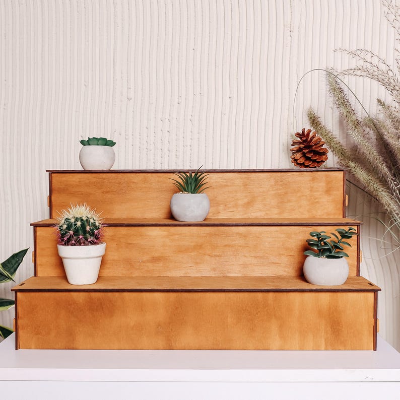 Pode incluir: Unidade de prateleira de madeira de tr&ecirc;s n&iacute;veis com acabamento em madeira natural. A unidade de prateleira &eacute; decorada com plantas em vasos, incluindo um cacto, uma suculenta e uma pequena &aacute;rvore.
