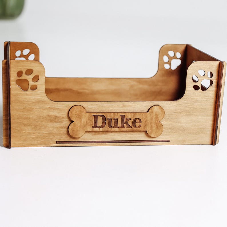 May include: A wooden dog bed with a bone-shaped cutout and the name "Duke" engraved on it. The bed has paw print cutouts on the sides.