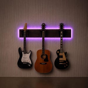 Può includere: Un supporto da parete per chitarre in legno scuro con illuminazione a LED viola integrata. Tre chitarre sono appese al supporto: una chitarra elettrica nera con battipenna bianco, una chitarra acustica in legno naturale e una chitarra elettrica nera.