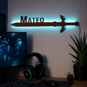 Può includere: Una lampada da parete a forma di spada in legno con il nome "MATEO" illuminato di blu. La spada ha un design dettagliato ed è montata su una parete bianca strutturata. Sono visibili un monitor del computer, cuffie e una pianta.