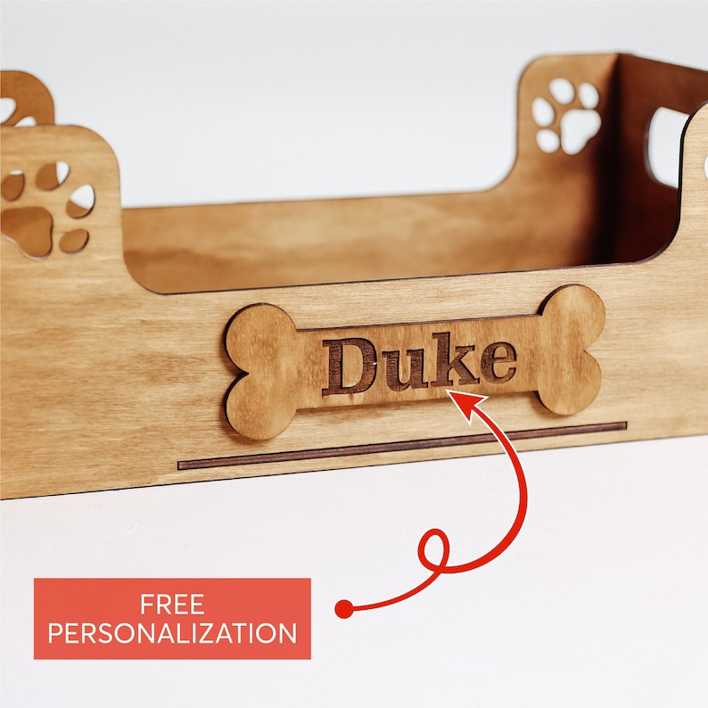 May include: Wooden dog bed with a bone-shaped plaque that says "Duke" on it. The bed is made of natural wood and has a paw print design on the side. Free personalization is offered.