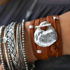 May include: A brown leather wrap bracelet with a silver charm featuring a peacock feather and the word "fun".
