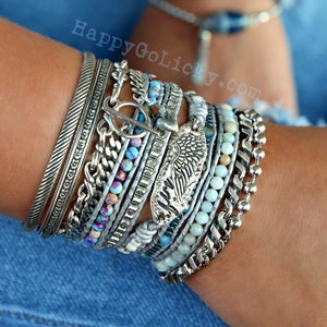 May include: A stack of silver bracelets with a variety of textures and styles, including a chain link bracelet, a beaded bracelet, and a bracelet with a silver wing charm. The bracelets are layered on a person's wrist.