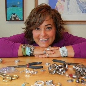 Può includere: Una donna con una camicia viola e anelli d'argento è seduta a un tavolo con una varietà di anelli d'argento, orecchini e altri gioielli.