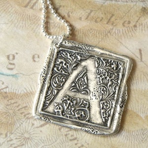 Op de afbeelding: Zilveren ketting met hanger met een vierkante bedel met een sierlijke letter A. De bedel heeft een gedetailleerd bloemmotief.