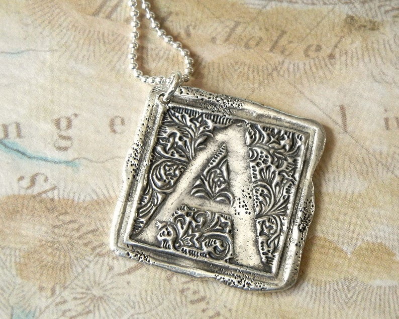Op de afbeelding: Zilveren ketting met hanger met een vierkante bedel met een sierlijke letter A. De bedel heeft een gedetailleerd bloemmotief.