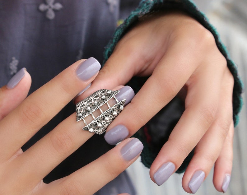 May include: A silver ring with a geometric design. The ring is worn on a finger with light grey nail polish.