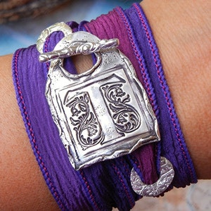 May include: A purple and burgundy silk ribbon bracelet with a silver charm featuring an ornate design. The charm has a hook closure.