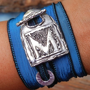 Puede incluir: Un brazalete de cinta azul y negro con un dije de plata con la letra "M" en una fuente decorativa.