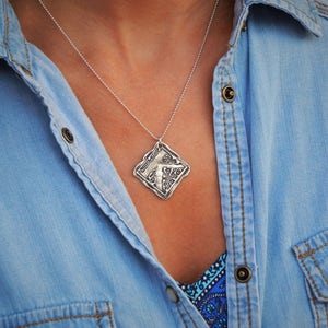 Op de afbeelding: Een zilveren ketting met een vierkante hanger met een gestileerde letter 'K'. De ketting wordt gedragen door iemand die een blauw denim shirt draagt.