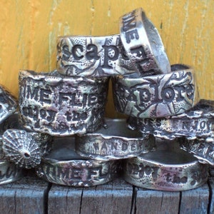 Puede incluir: Una pila de anillos de plata con diferentes palabras y diseños grabados en ellos. Las palabras en los anillos incluyen "Time Flies", "Wander", "Explore", "Escape" y "Remember".