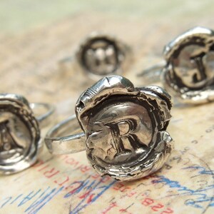 May include: Four silver rings with a wax seal design. Each ring features a different letter, R, J, M, and I, embossed in the center of the seal.