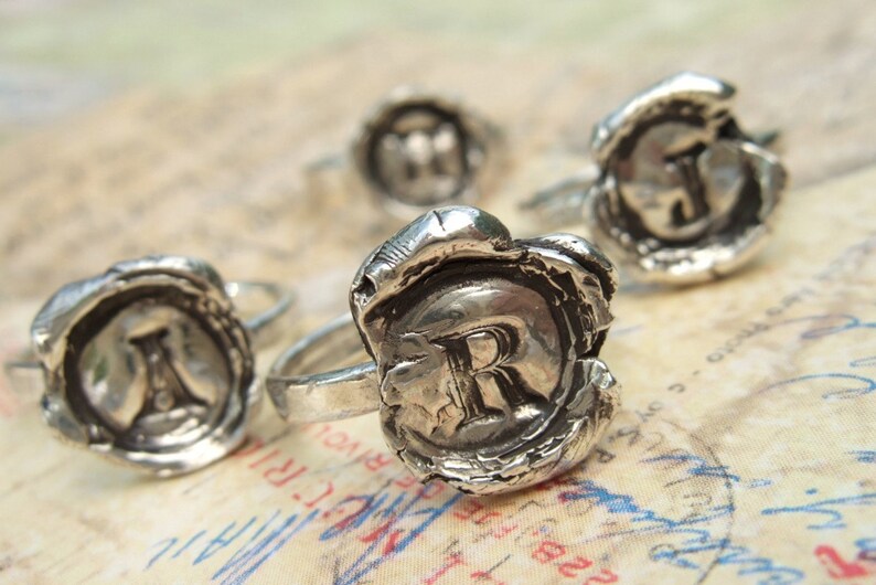 May include: Four silver rings with a wax seal design. Each ring features a different letter, R, J, M, and I, embossed in the center of the seal.