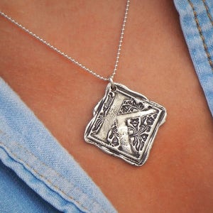 Op de afbeelding: Een zilveren ketting met een vierkante hanger met een reli&euml;fletter K omringd door een bloemmotief.