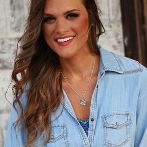 May include: A woman with long brown hair wearing a light blue denim shirt with a silver necklace and a pendant.