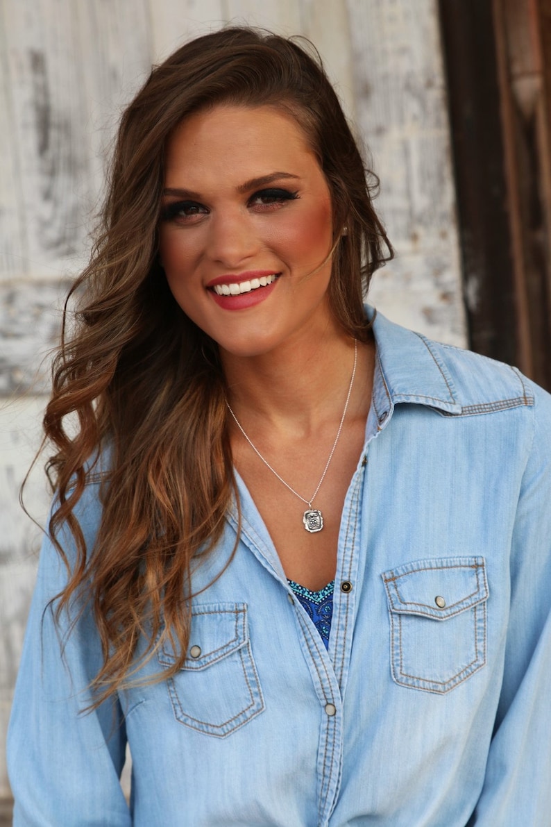 May include: A woman with long brown hair wearing a light blue denim shirt with a silver necklace and a pendant.