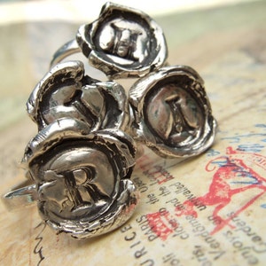 May include: Three silver rings with wax seal designs. Each ring features a different letter, J, R, and A, in a raised, stylized font.