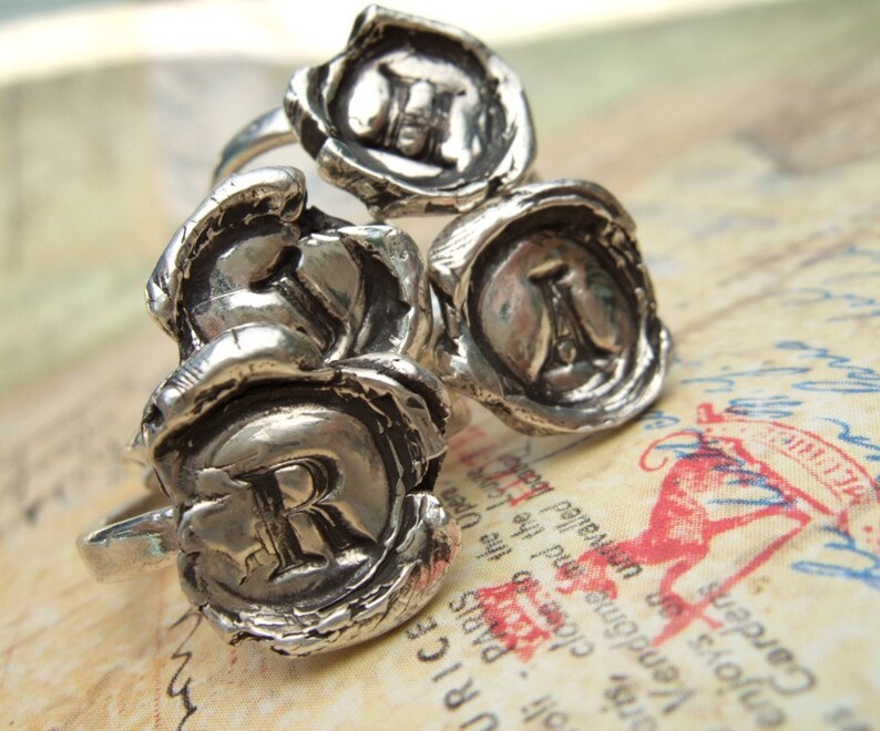 May include: Three silver rings with wax seal designs. Each ring features a different letter, J, R, and A, in a raised, stylized font.