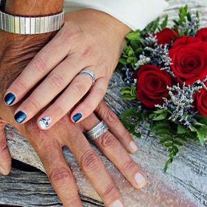 May include: A close-up of two hands with wedding bands on their ring fingers. The hands are resting on a piece of weathered wood. A bouquet of red roses with green leaves is in the background.