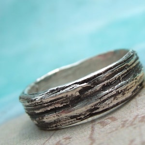 May include: A silver ring with a textured, wood-like design. The ring is wide and has a rustic, organic look.