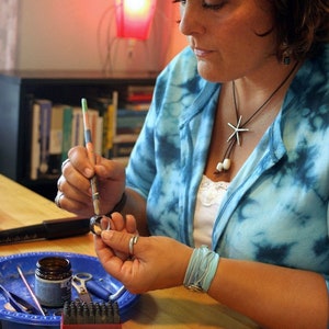 K&ouml;nnte beinhalten: Eine Frau arbeitet an einem Schmuckprojekt. Sie verwendet einen kleinen Pinsel, um einen Versiegler auf einen Metallring aufzutragen. Der Ring liegt auf einem blauen Tablett mit verschiedenen Werkzeugen und Materialien. Die Frau tr&auml;gt ein blaues und wei&szlig;es Batik-Shirt und eine silberne Halskette mit einem Seestern-Anh&auml;nger.
