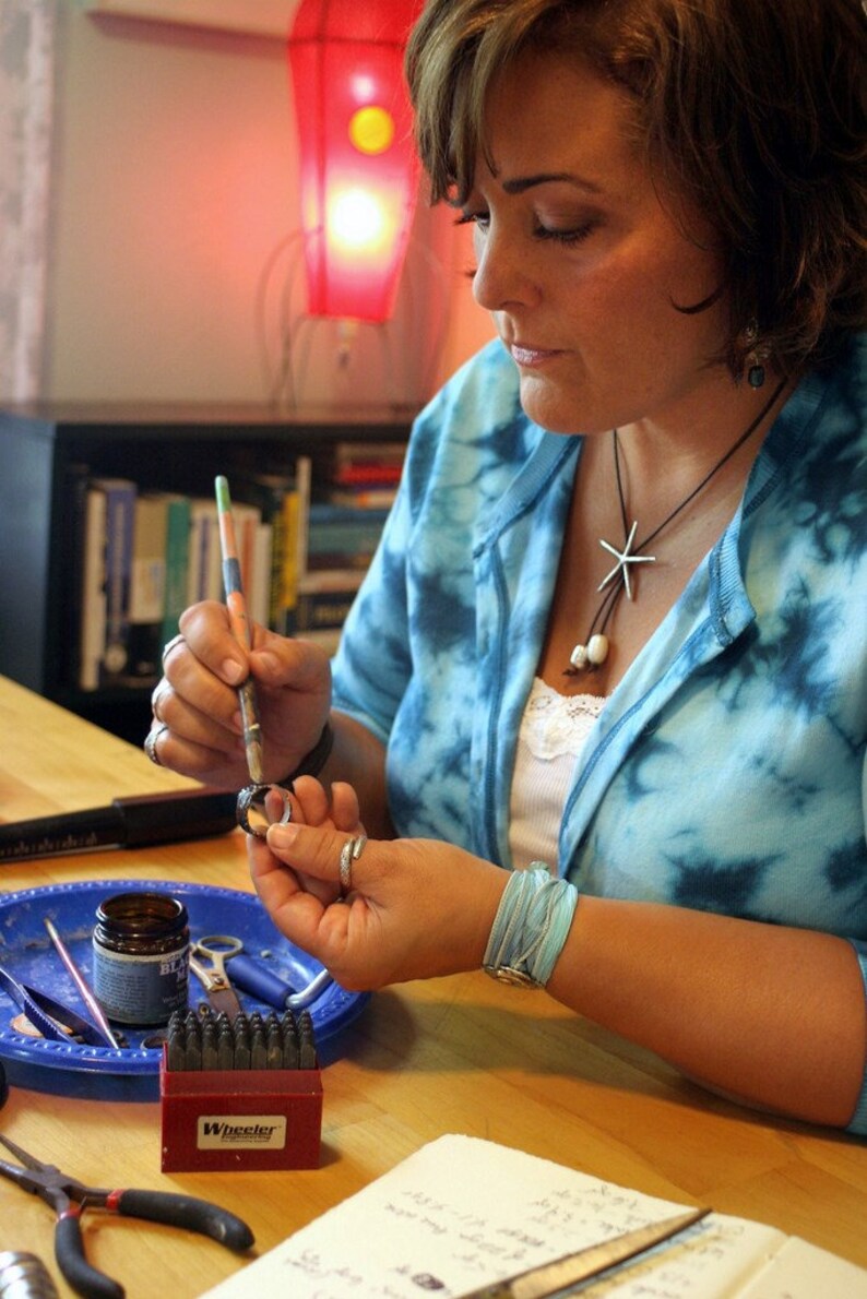 K&ouml;nnte beinhalten: Eine Frau arbeitet an einem Schmuckprojekt. Sie verwendet einen kleinen Pinsel, um einen Versiegler auf einen Metallring aufzutragen. Der Ring liegt auf einem blauen Tablett mit verschiedenen Werkzeugen und Materialien. Die Frau tr&auml;gt ein blaues und wei&szlig;es Batik-Shirt und eine silberne Halskette mit einem Seestern-Anh&auml;nger.