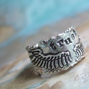 May include: Silver ring with a textured design featuring a pair of wings and the word "TRUST" embossed on the band.