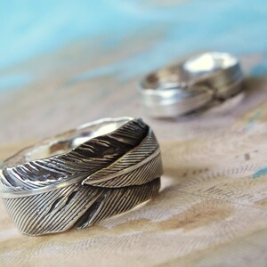 May include: Two silver rings with a feather design. The rings are on a light brown surface with a blue background.