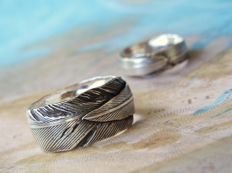 May include: Two silver rings with a feather design. The rings are on a light brown surface with a blue background.