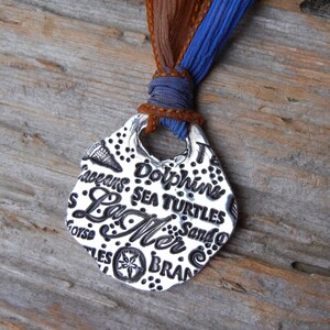 May include: A silver pendant necklace with a seashell-shaped charm. The charm is engraved with the words "Oceans", "Dolphins", "Sea Turtles", "La Mer", "Sand", and "Bran", along with a starfish design. The necklace is attached to a blue and brown ribbon.
