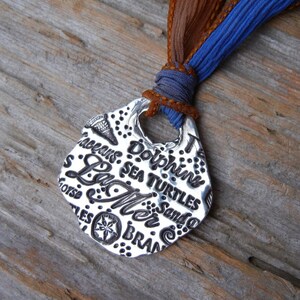 May include: Silver pendant necklace with a seashell design. The pendant features the words "Sea Turtles", "Sand", "Sea", "La Mer", "Dolphins", "Shells", "Ocean", and "Brawn". The necklace is attached to a brown and blue ribbon.