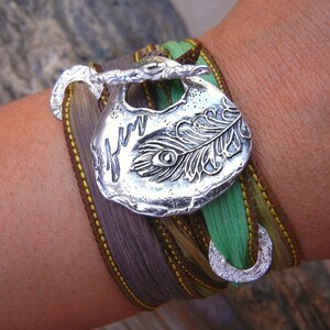Puede incluir: Pulsera plateada con un colgante grande y ornamentado con un diseño de pluma de pavo real. La pulsera tiene múltiples bandas de tela en tonos verdes, marrones y grises, con costuras amarillas. Cierre plateado.