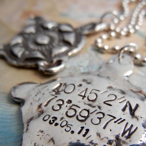 May include: Silver necklace with two pendants. One pendant is a stylized flower design. The other pendant is a silver heart with the coordinates 40.4522°N 73.5937°W and the date 03.05.11 engraved on it.