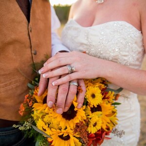 May include: A close-up of a couple's hands with wedding rings, holding a bouquet of yellow sunflowers and orange flowers.