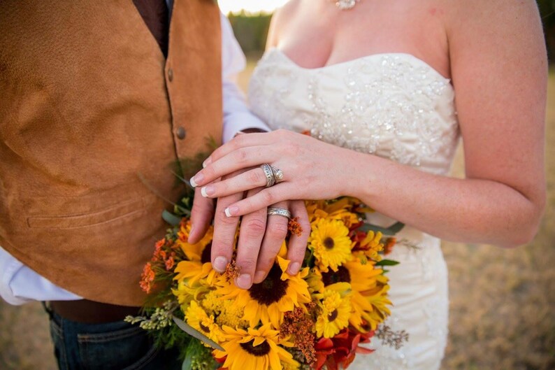 May include: A close-up of a couple's hands with wedding rings, holding a bouquet of yellow sunflowers and orange flowers.