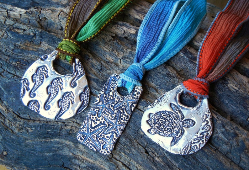 May include: Three silver pendants with different designs: a seahorse, a starfish, and a turtle. Each pendant is attached to a colorful ribbon.