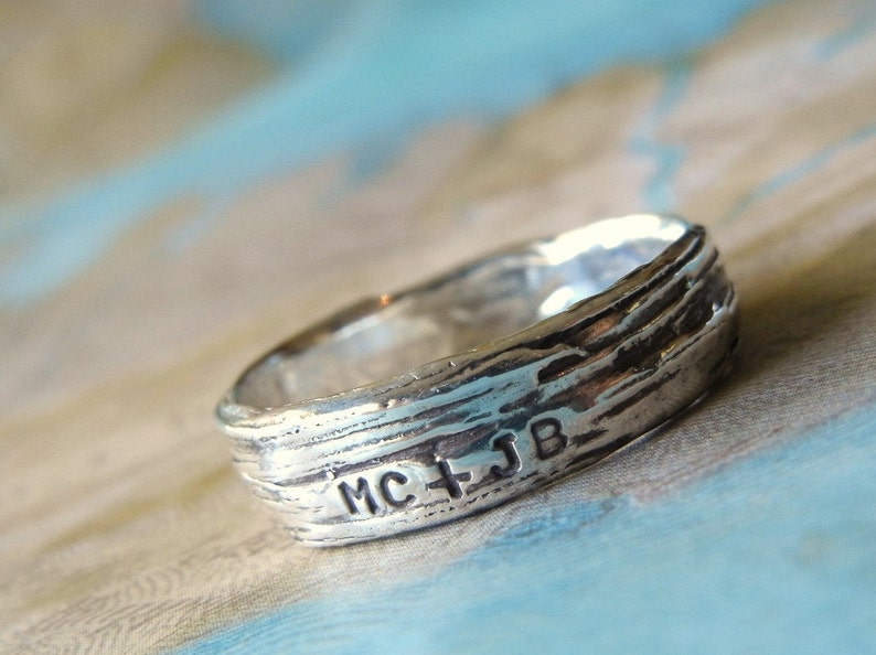 May include: A silver ring with a textured, wood-like design. The ring is engraved with the initials "MC + JB".