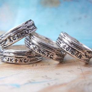 May include: Four silver rings with engraved text. The rings are stacked on top of each other. The text on the rings reads "ACH+PL", "JK+NC", "ER+TC", and "SO+BR".