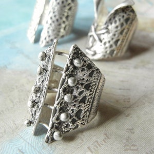 May include: A silver ring with a textured design and a cage-like structure. The ring has a geometric shape and is adorned with small, round beads.