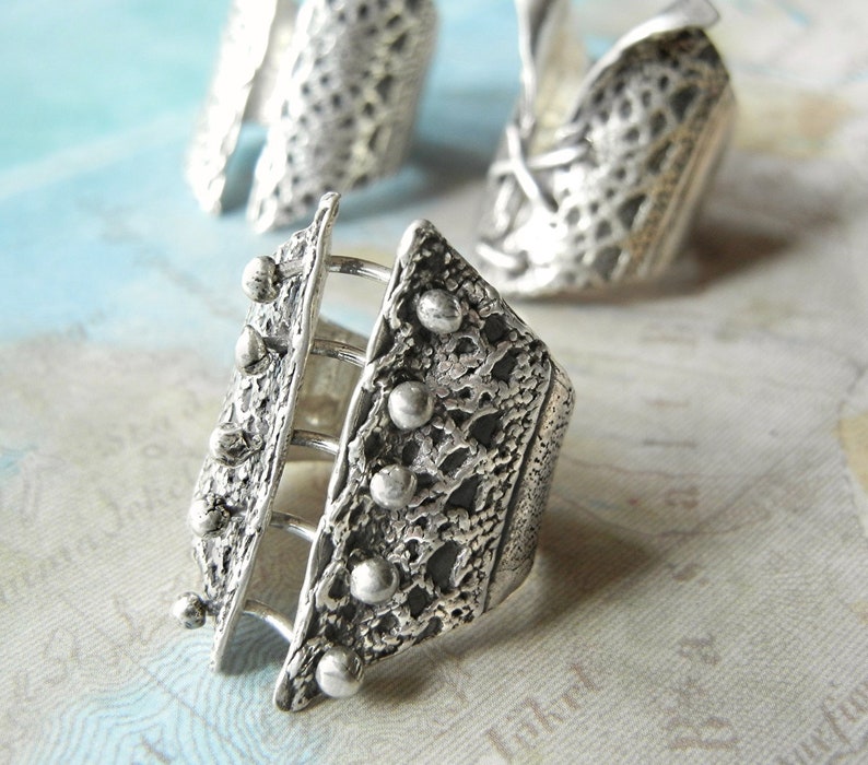 May include: A silver ring with a textured design and a cage-like structure. The ring has a geometric shape and is adorned with small, round beads.