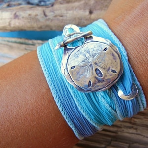 May include: A blue and turquoise fabric bracelet with a silver sand dollar charm. The bracelet is wrapped around a wrist.
