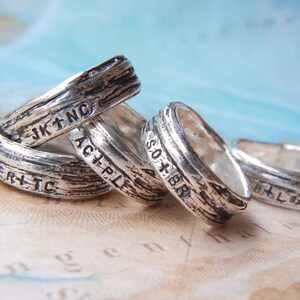 May include: Four silver rings with engraved text, including "Arctic", "Pacific", "South Bend", and "JK+NC". The rings are stacked on top of each other and have a textured, rustic finish.
