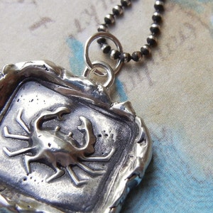 May include: A silver pendant necklace featuring a detailed crab design. The pendant is set in a square frame with a textured, aged finish. The chain is made of silver beads.